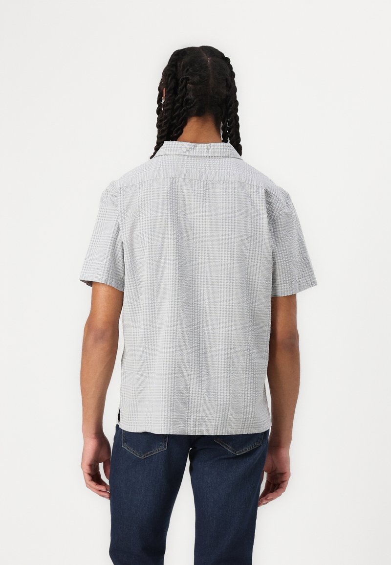 Chemise à manches courtes en gris clair, avec un motif à carreaux texturé. Col classique et coupe décontractée. Portée avec un jean foncé.