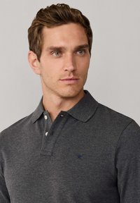 Polo gris foncé en tissu doux avec un col, trois boutons et un petit logo brodé bleu sur la poitrine.