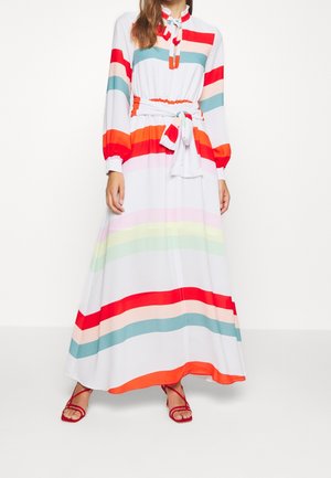Kobieta w długiej białej sukience z poziomymi czerwonymi, pomarańczowymi, różowymi, zielonymi i niebieskimi paskami, z talią podkreśloną paskiem, oraz w czerwonych sandałach na paski.