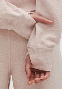 Bluza lungă cu mâneci, de culoare roz deschis, prezentând un design texturat de tip waffle-knit, cu detalii cu orificii pentru degete și o croială lejeră în jurul încheieturilor.