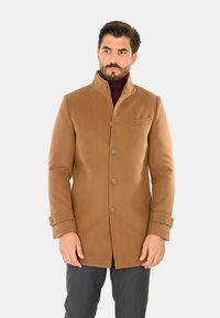 Cappotto di lana marrone con colletto alto, chiusura anteriore con bottoni, tasche laterali e taschino sul petto. Il modello indossa un dolcevita scuro sotto.