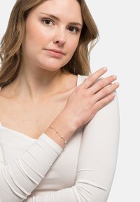 Bracciale d'oro con piccole perle rotonde, indossato al polso. Il modello ha la pelle chiara e indossa un top aderente bianco a maniche lunghe.