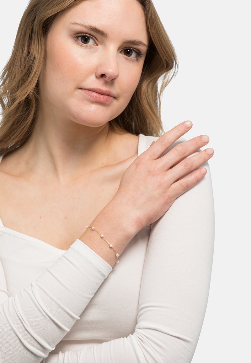 Bracciale d'oro con piccole perle rotonde, indossato al polso. Il modello ha la pelle chiara e indossa un top aderente bianco a maniche lunghe.