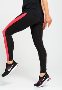 Černé sportovní legíny s růžovým postranním pruhem, se štíhlým střihem a hladkou texturou. Sláčeny s černými a růžovými běžeckými botami.