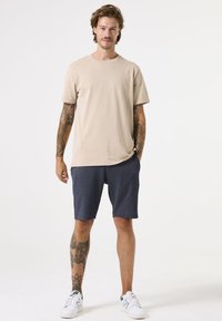Beige katoenen t-shirt met korte mouwen, gecombineerd met marineblauwe shorts van een gestructureerde stof, compleet gemaakt met witte sneakers met groene accenten.