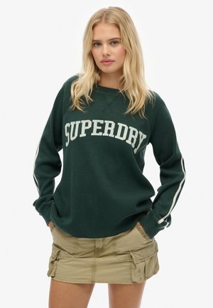 Green, ribbed long-sleeve sweatshirt featuring "SUPERDRY" logo in white, paired with a beige cargo skirt with side pockets.