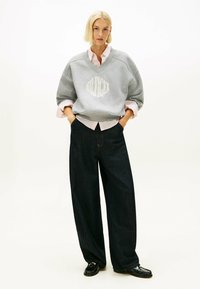 Felpa grigia con scollo a V e logo, sovrapposta a una maglietta a righe rosa chiaro. Jeans neri a gamba larga e scarpe nere completano l'outfit.