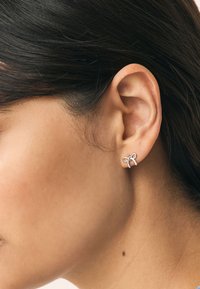 Srebrna naušnica u obliku mašne sa glatkom, poliranom površinom. Tanki naglasci tvore ukrasni čvor, pričvršćen za mali klin.