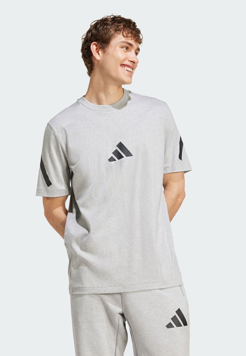 Young man smiling, wearing light gray Adidas t-shirt and matching pants with black logo on chest, sleeves, and leg.