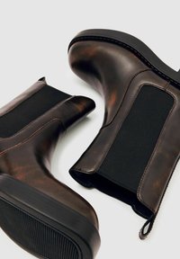 Rudi odiniai aukšti botai su elastiniais šonų intarpais, apvalia nosimi ir gaubtuotomis gumos padais, demonstruojantys elegantišką dizainą ir kontrastingas medžiagas.