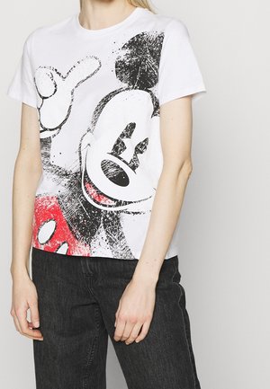 Kobieta nosząca białą koszulkę z dużym, postarzanym nadrukiem Myszki Miki w czerni i czerwieni, zestawioną z czarnymi jeansami na białym tle.