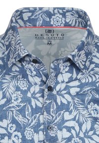Korte mouwen shirt van blauwe stof met witte bloem- en blaadjespatronen. Het heeft een klassieke kraag en twee zwarte knopen aan de voorkant.
