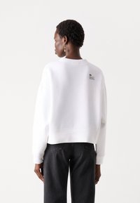 White sweatshirt featuring a crew neck, relaxed fit, and ribbed cuffs, with a small logo patch on the back. Paired with black jeans.