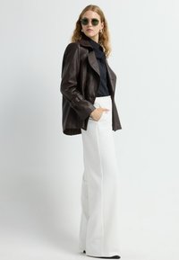 Giacca in pelle marrone, camicia nera con bottoni, pantaloni bianchi a gamba larga, occhiali da sole tondi verdi, orecchini in argento, tessuto testurizzato, vestibilità sartoriale.