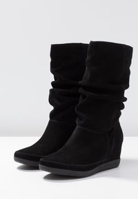 Bottes compensées en daim noir avec une tige froncée, présentant un bout pointu et une surface texturée, contrastant avec une semelle lisse noire.