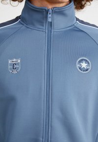 Chaqueta azul con cremallera fabricada en tejido suave. Presenta logotipos bordados: un escudo y una estrella circular en el pecho. Dispone de franjas laterales en contraste.