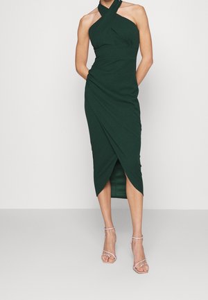 Femme portant une robe portefeuille midi sans manches vert foncé avec un col halter croisé et des sandales à talons hauts avec lanières.