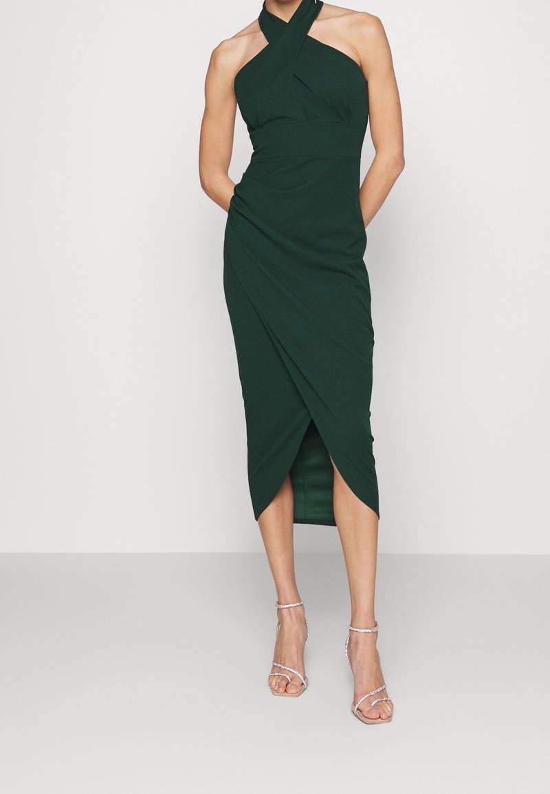 Femme portant une robe portefeuille midi sans manches vert foncé avec un col halter croisé et des sandales à talons hauts avec lanières.
