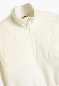 Pull-over zippé crème clair fabriqué en tissu matelassé, avec une section supérieure texturée et une partie inférieure lisse.
