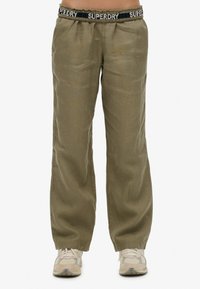 Pantalons olive vert, amples, avec une ceinture élastique affichant "SUPERDRY" en lettres blanches. Le matériau semble doux avec une texture minimale.