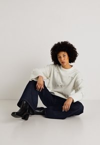 Balta mezgimo megztinis su kaspino akcentais, derinamas su tamsiomis plačiomis džinsomis ir juodais auliniais batais. Sklandus audinys ir laisvas fasonas.