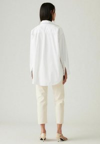 Camisa blanca de algodón de corte oversize con dobladillo curvado, combinada con jeans cortos de color blanco sucio y mules de tacón beige. Diseño simple y limpio.