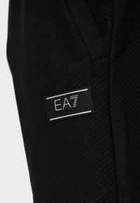 Ύφασμα μαύρου αθλητικού ενδύματος με υφή διάτρητου πλέγματος, με προσαρτημένη ορθογώνια ετικέτα EA7 Emporio Armani.