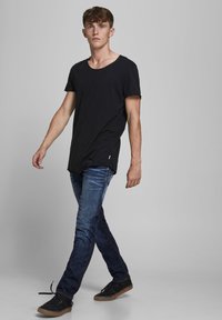 Joven caminando, vistiendo una camiseta negra, jeans azules y zapatillas negras, frente a un fondo gris claro liso.