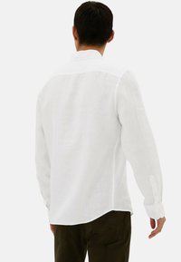 Camisa de lino blanca de manga larga con cuello abotonado, que presenta un dobladillo curvado y detalles de puños abotonados. Textura sutil y ligera.