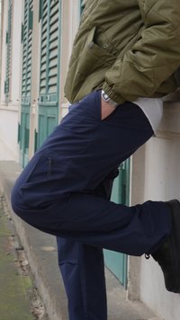 Άτομο που ακουμπάει σε έναν τοίχο με το ένα πόδι λυγισμένο, φορώντας πράσινο μπουφάν, μπλε παντελόνι, μαύρα παπούτσια και ασημένιο ρολόι χειρός.