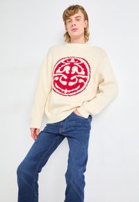 Jeune personne portant un pull crème avec un grand motif circulaire rouge et un jean bleu, posant avec une main dans la poche sur fond blanc.