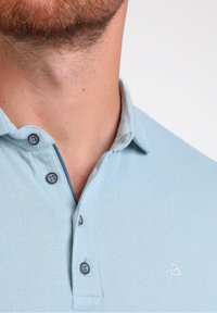 Šviesiai mėlynas polo marškinėliai su tekstūruota paviršiumi, trimis mygtukais ant apykaklės ir maža logotipo emblema ant krūtinės.