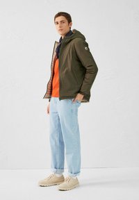 Veste légère vert olive avec capuche, fermeture éclair, doublure beige, chemise orange, jean bleu clair et baskets beiges.