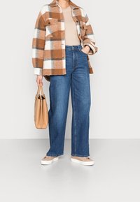 Person med brun och vit rutig jacka, beige topp, blå vida byxor, beige sneakers, som håller en beige handväska vid sidan.