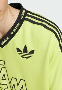 Žalias sportinis džersis su juodais akcentais, turintis raukšlėtą apykaklę ir tris juodas juosteles ant pečių. Apima juodą logotipą.