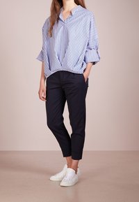 Gestreifte blaue und weiße Oversized-Bluse mit V-Ausschnitt und weiten Ärmeln, kombiniert mit maßgeschneiderten navyblauen Hosen und weißen Sneakern.