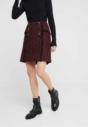 Gonna in tweed rosso con bottoni neri e bordi sfrangiati, abbinata a stivaletti neri in pelle con fibbie dorate e suole testurizzate.