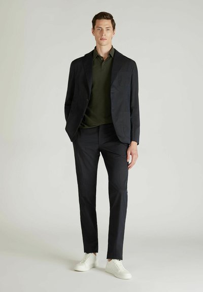 Blazer azul marino con un único bolsillo, combinado con una polo verde oscuro y pantalones negros, complementado con zapatillas blancas. Tela suave, corte ajustado.