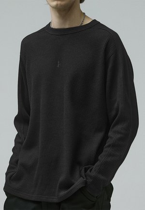 Black long-sleeve sweater made of textured fabric. Features a ribbed crew neckline and a subtle logo embroidered on the chest.