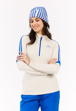 Beige lynlåsbluse med blå detaljer og ærmer, der har en høj krave. Strikket hvid og blåstribet hue. Glat stofstruktur.