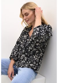 Sorte blomsterbluse med pufærmer, dyb V-hals og hvide blomster mønstre. Parret med lyseblå jeans, siddende op ad en hvid væg.