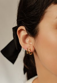 Boucles d'oreilles en or avec une finition polie, portées sur le lobe de l'oreille, mettant en valeur trois anneaux empilés et un ruban sombre partiellement visible dans les cheveux.