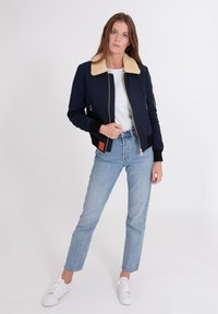 Marine bomberjack met een nepbont kraag, ritssluiting en oranje label; gecombineerd met lichtblauwe jeans en witte sneakers.