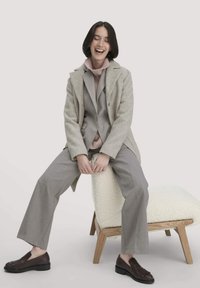 Hellgrauer Wollmischmantel über einem grauen Anzug, kombiniert mit einem pinkfarbenen Strickrollkragenpullover und braunen Lederschuhen, sitzend auf einem strukturierten Hocker.