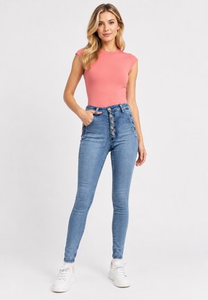 Kvinde med langt blondt hår iført en ærmeløs koral top, højtaljede blå skinny jeans med knapper foran og hvide sneakers, stående med hånden i lommen.