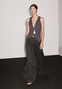 Pull en tricot à col en V profond gris foncé, associé à un pantalon à pinstripes noir et large. Le modèle porte un long collier en argent et des sandales noires.