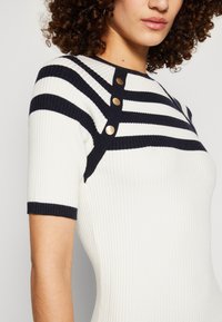 Haut en maille côtelée à rayures blanches et bleu marine, avec boutons aux épaules et manches courtes. Tissu texturé avec des couleurs contrastantes et des détails soignés.