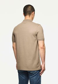 Man gezien vanuit de rug in een beige polo met korte mouwen en blauwe jeans tegen een effen lichte achtergrond.