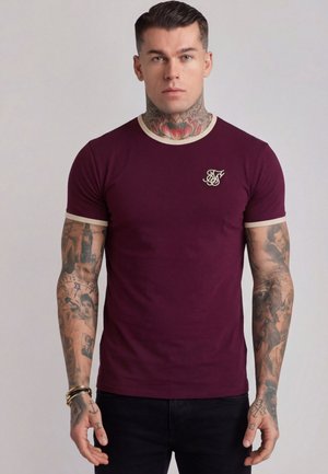 Man with neck and arm tattoos wears a fitted maroon T-shirt with beige trim and monogram logo, standing against a plain background.