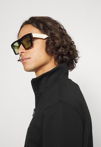 Zwarte oversized zonnebril met geel getinte lenzen, voorzien van dikke zwarte monturen en crèmekleurige armpjes, gedragen met een zwarte ritsjack.
